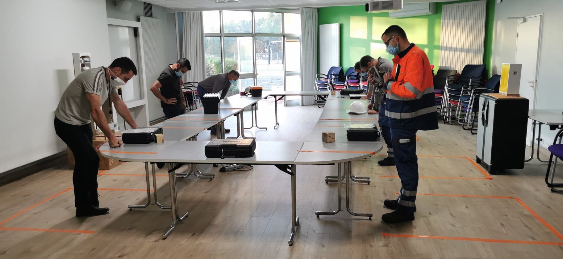 Photo d'un groupe de personnes en train de lire des consignes devant une table. Les personnes sont dispersées sur une grande table pour respecter les gestes barrières.